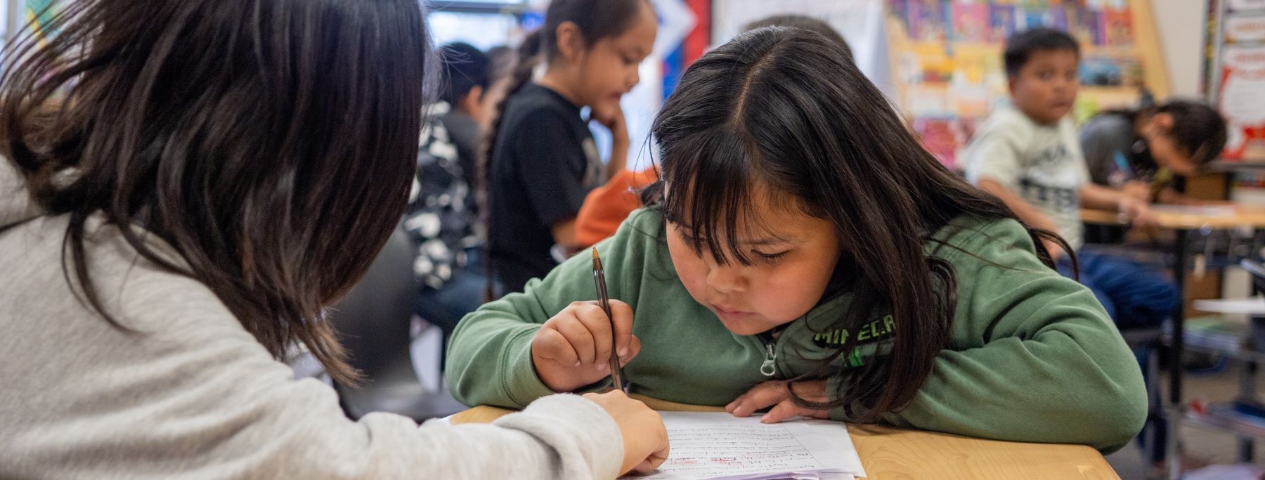 A young girl sits at a table, focused on writing in her notebook with a pencil in a busy classroom..