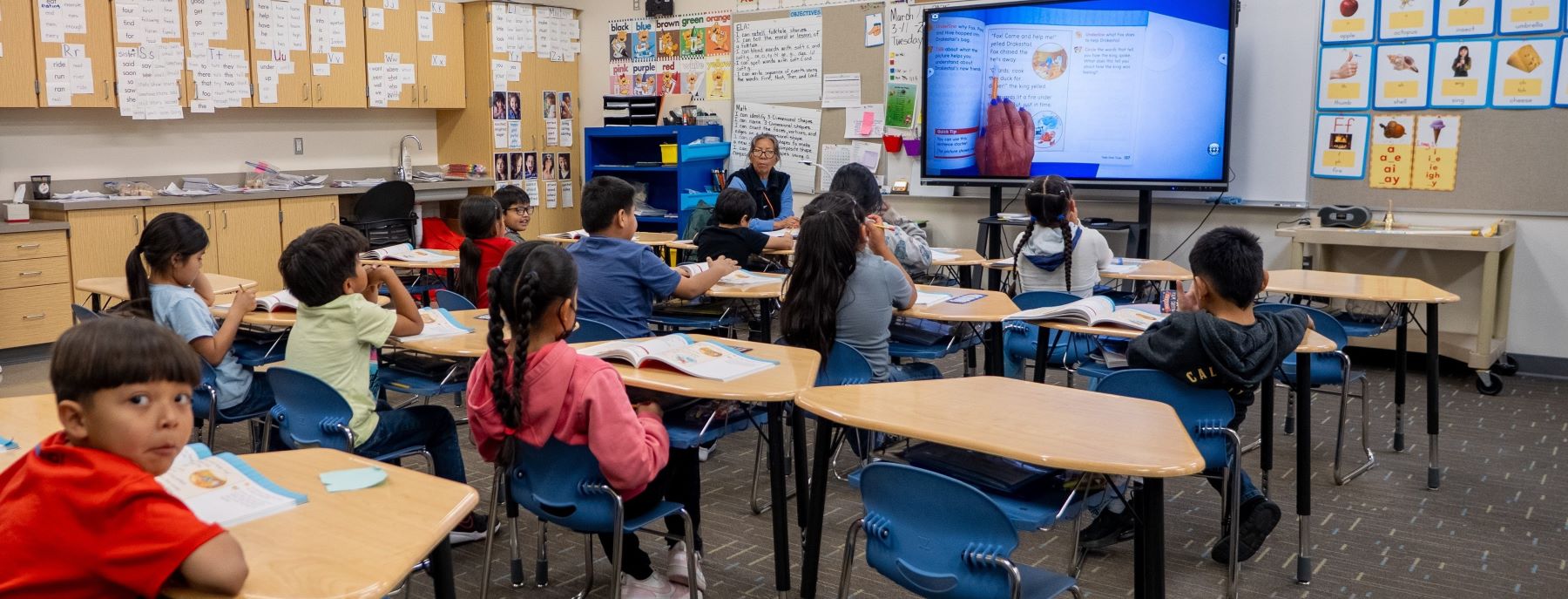Inside a classroom, children are seated at desks while a teacher at the front, leading the lesson.